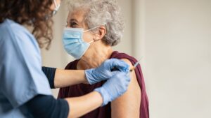 pessoa de terceira idade usando máscara recebe vacina no braço, aplicada por uma profissional de saúde com luvas e máscara.
