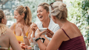 Grupo de mulheres maduras sorrindo e conversando alegremente ao redor de uma mesa ao ar livre, com frutas e bebidas à frente — celebrando a vida, a amizade e a sorte de estarem juntas.