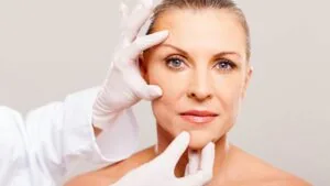 Mulher madura sendo examinada por profissional de saúde com luvas, em avaliação prévia para cirurgia plástica facial.