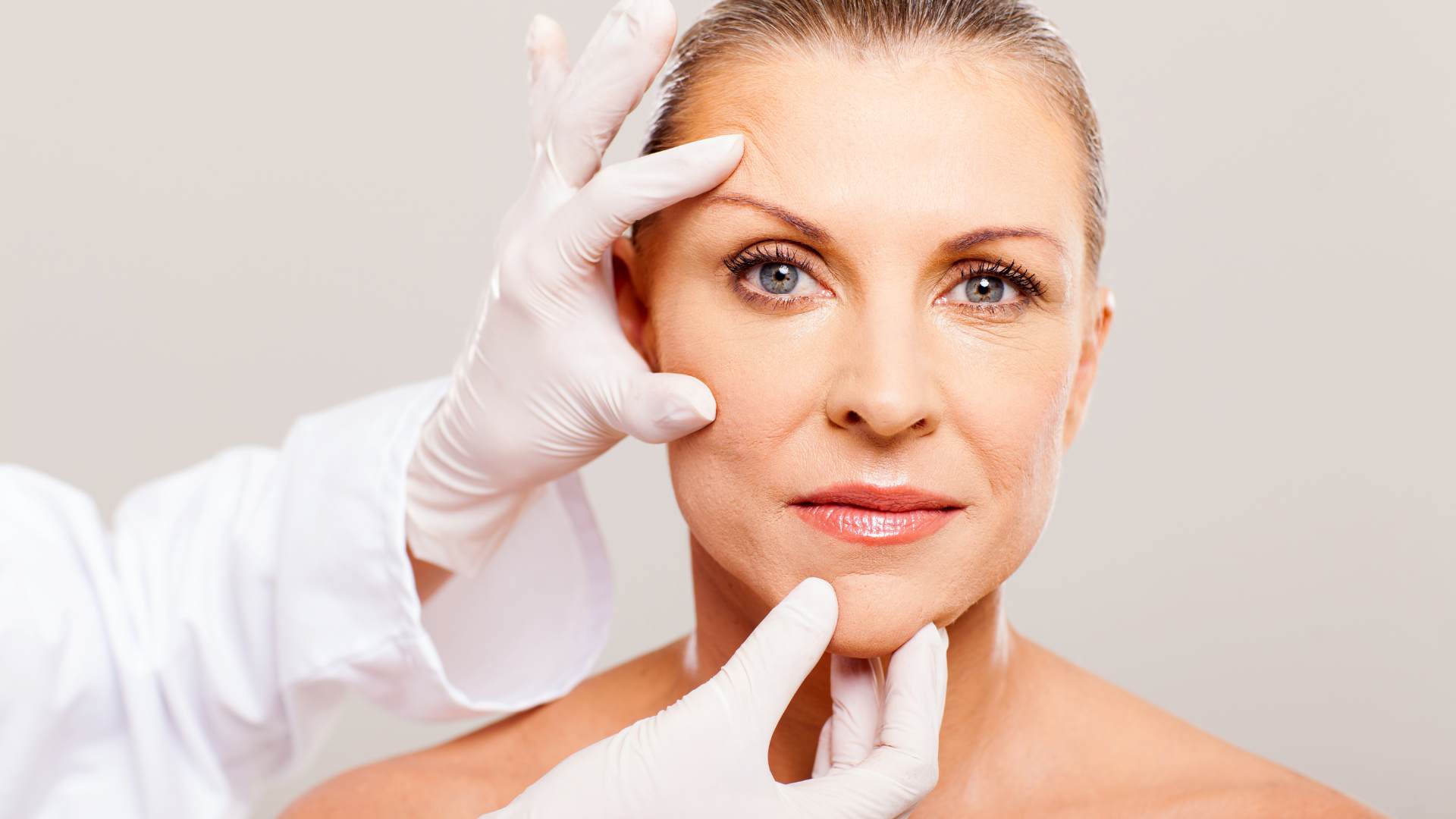 Mulher madura sendo examinada por profissional de saúde com luvas, em avaliação prévia para cirurgia plástica facial.
