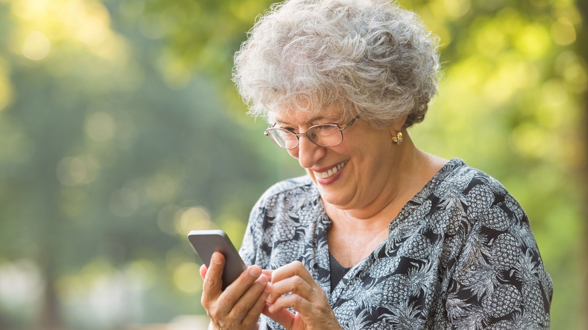 Mulher 50+ sorrindo enquanto usa o celular ao ar livre.