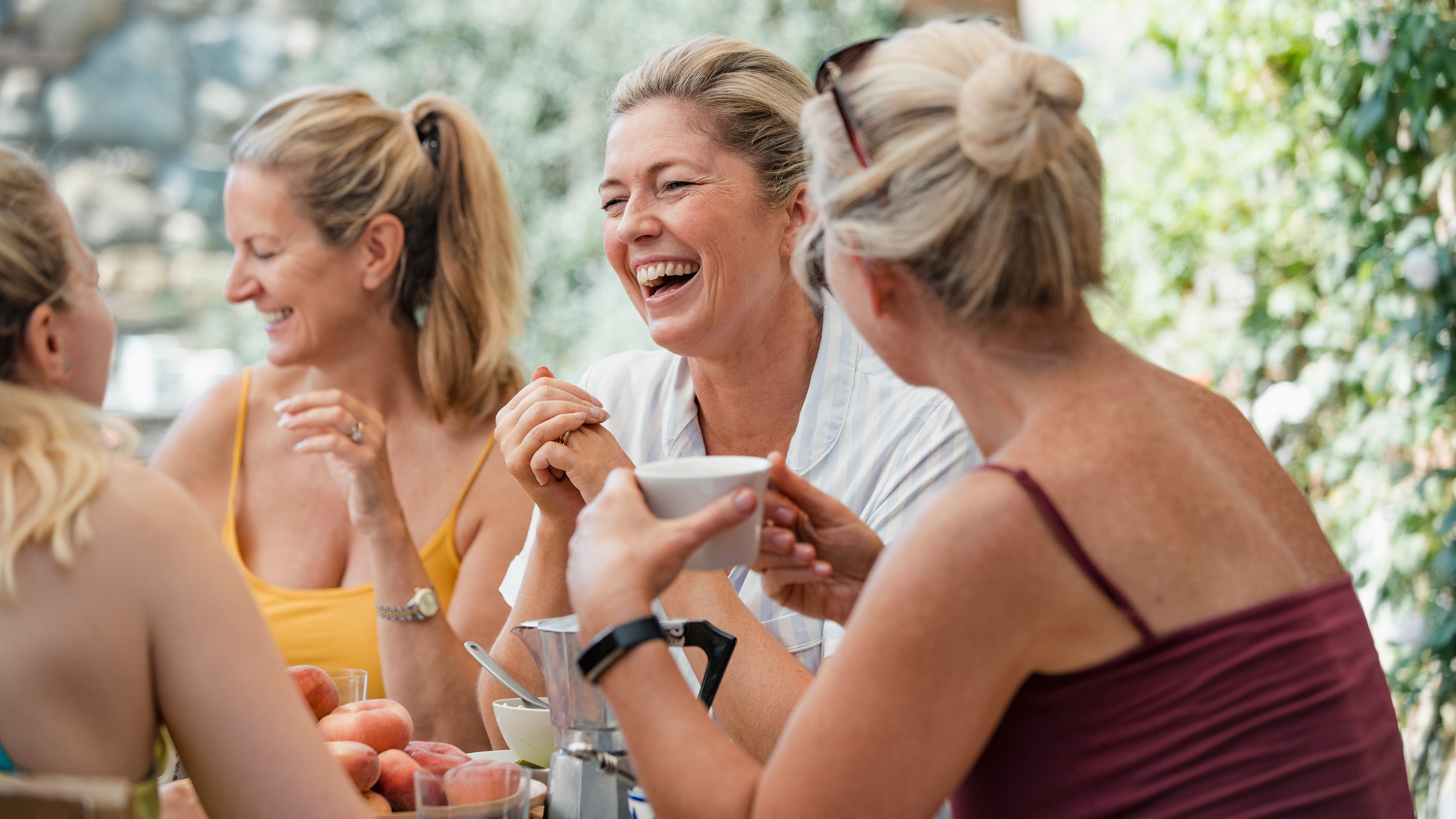 Grupo de mulheres maduras sorrindo e conversando alegremente ao redor de uma mesa ao ar livre, com frutas e bebidas à frente — celebrando a vida, a amizade e a sorte de estarem juntas.